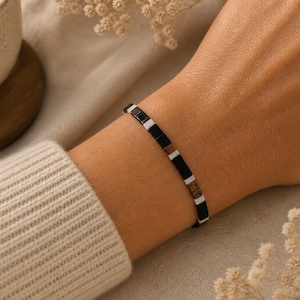 Puede incluir: Una pulsera minimalista con pequeñas cuentas rectangulares en negro, blanco y marrón. La pulsera se lleva en la muñeca, con una manga de suéter de tonos neutros visible. El fondo incluye una taza y flores secas.
