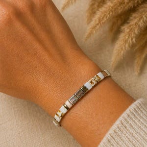 Op de afbeelding: Een armband met vierkante kralen in wit, goud en bruin. De armband wordt om een pols gedragen, tegen een neutrale achtergrond met gedroogde planten. De armband heeft een mix van witte, gouden en bruine kralen.
