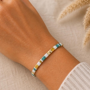 Puede incluir: Un brazalete de cuentas con cuentas rectangulares turquesas, blancas y doradas. El brazalete se muestra en una muñeca. El fondo es neutro, con plantas secas. Una joya delicada.