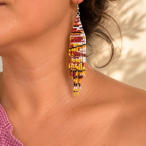 Peut inclure: Une longue boucle d'oreille pendante composée de petites perles de rocaille formant un motif géométrique. Le perlage présente une palette de couleurs vives rouge, jaune, blanche et noire, disposées en bandes horizontales effilées.