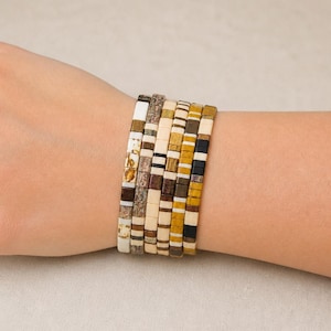 Op de afbeelding: Een stapel van zes armbanden met kralen in verschillende kleuren en patronen. De armbanden hebben rechthoekige kralen in tinten wit, bruin, beige en zwart. De armbanden worden om een pols gedragen.