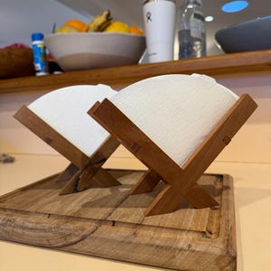 Può includere: Due portafiltri per caffè in legno con filtri di carta bianchi, appoggiati su un tagliere di legno. I supporti sono in legno marrone scuro e hanno un design a forma di X. I filtri sono bianchi e triangolari.