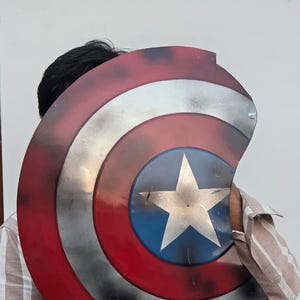 May include: A Captain America shield prop with a red, white, and blue color scheme. The shield features a white star on a blue circle, surrounded by white and red stripes. The prop is held by a person.