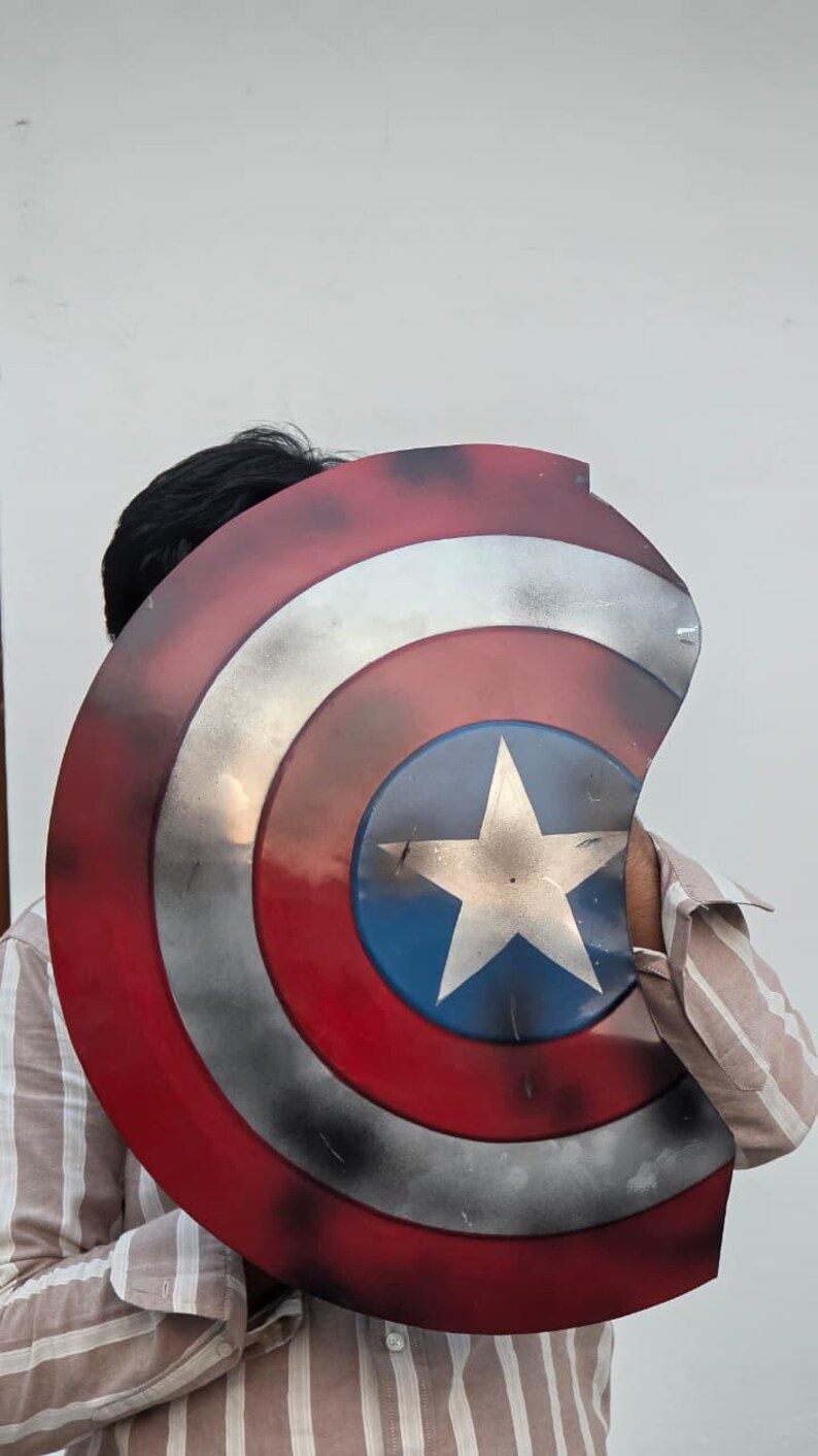 May include: A Captain America shield prop with a red, white, and blue color scheme. The shield features a white star on a blue circle, surrounded by white and red stripes. The prop is held by a person.