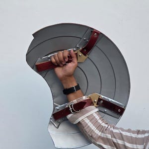 May include: A gray, shield-shaped prop with red and gold accents. The prop has a circular design with radial lines and is held by a person. The shield has red leather straps and gold-colored metal hardware.