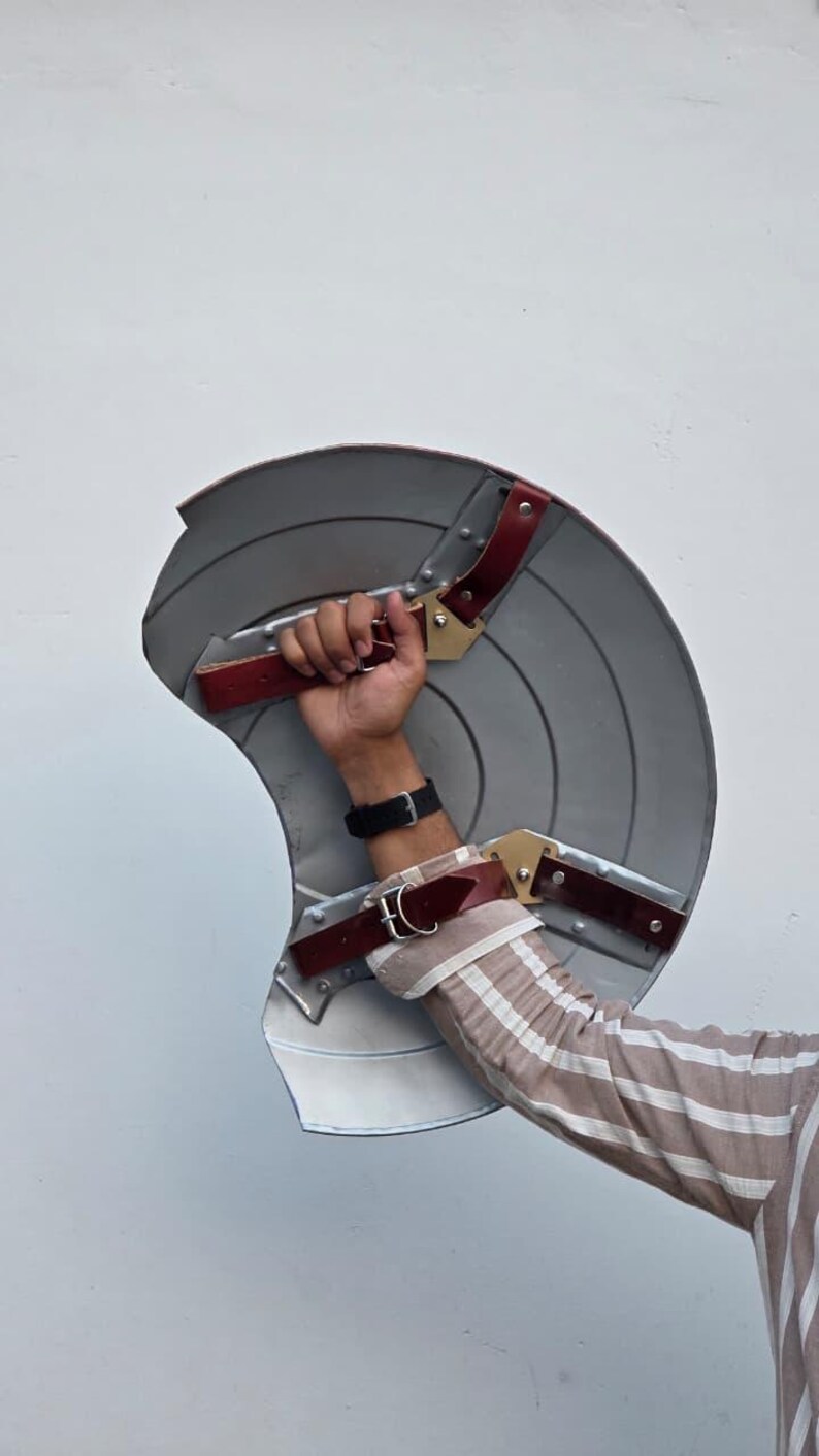 May include: A gray, shield-shaped prop with red and gold accents. The prop has a circular design with radial lines and is held by a person. The shield has red leather straps and gold-colored metal hardware.