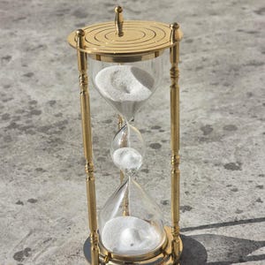 May include: A gold-colored hourglass with clear glass chambers and white sand. The hourglass has a decorative top and base, and is supported by four vertical gold-colored bars. The hourglass is approximately 6 inches tall.