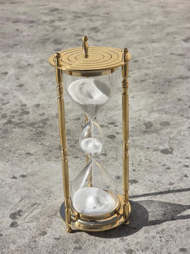 May include: A gold-colored hourglass with clear glass chambers and white sand. The hourglass has a decorative top and base, and is supported by four vertical gold-colored bars. The hourglass is approximately 6 inches tall.