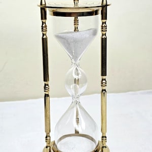 Peut inclure: Un sablier en laiton avec des chambres en verre transparent et une ampoule centrale. Le sablier est soutenu par un cadre en laiton avec des détails décoratifs. Le sable à l'intérieur est blanc. Le sablier mesure environ 18 cm de haut.