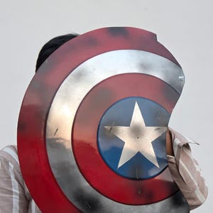May include: A Captain America shield prop. The shield features a red, white, and blue color scheme with a silver star in the center. The shield has a distressed, battle-worn appearance, with visible wear and tear.