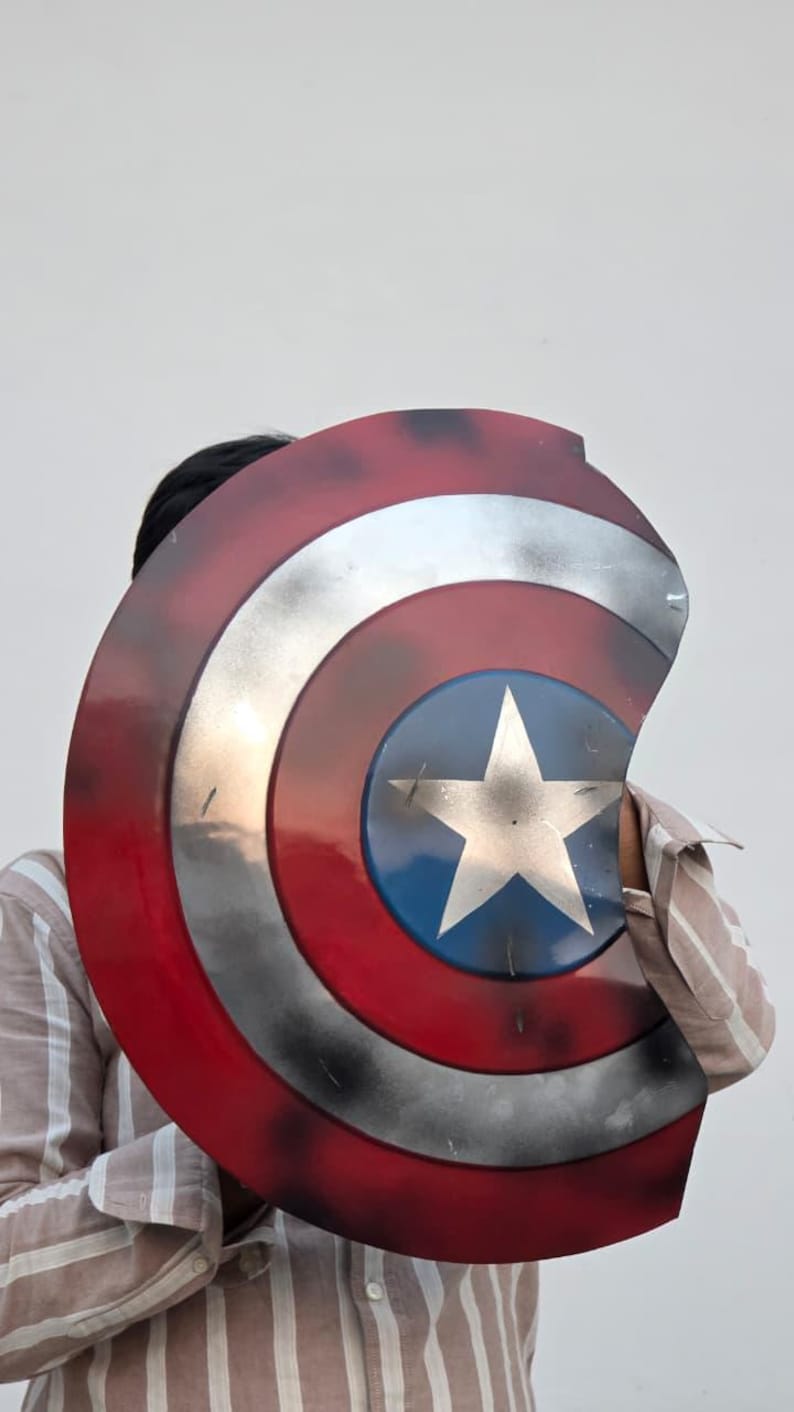 May include: A Captain America shield prop. The shield features a red, white, and blue color scheme with a silver star in the center. The shield has a distressed, battle-worn appearance, with visible wear and tear.