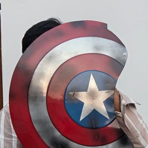 May include: A Captain America shield prop, featuring a red, white, and blue color scheme. The shield has a circular design with a white star on a blue circle at the center. The shield is made of metal and has a distressed finish.