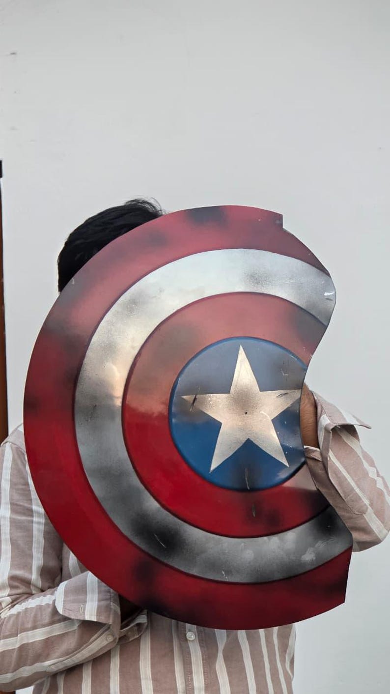 May include: A Captain America shield prop, featuring a red, white, and blue color scheme. The shield has a circular design with a white star on a blue circle at the center. The shield is made of metal and has a distressed finish.