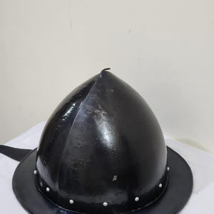 Könnte beinhalten: Ein schwarzer Metallhelm mit abgerundeter Oberseite und breitem Rand. Der Helm hat eine Reihe kleiner, gleichmäßig verteilter Nieten um den Rand. Die Oberfläche des Helms hat ein leicht strukturiertes Aussehen.