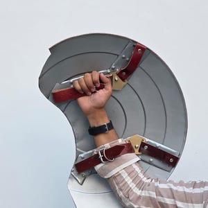 May include: A silver-colored shield with red and gold accents. The shield has a curved design with concentric circles and leather straps for gripping. A replica of a superhero shield.