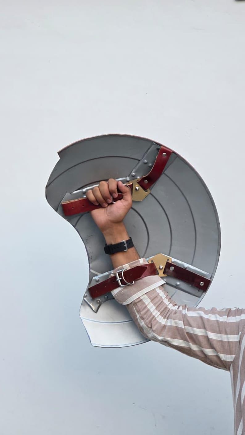 May include: A silver-colored shield with red and gold accents. The shield has a curved design with concentric circles and leather straps for gripping. A replica of a superhero shield.