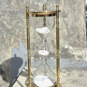 May include: A brass hourglass with clear glass chambers and white sand. The hourglass has a decorative, ornate design with a round base and top. The sand is flowing from the top chamber to the bottom chamber. The hourglass is a classic timekeeping device.