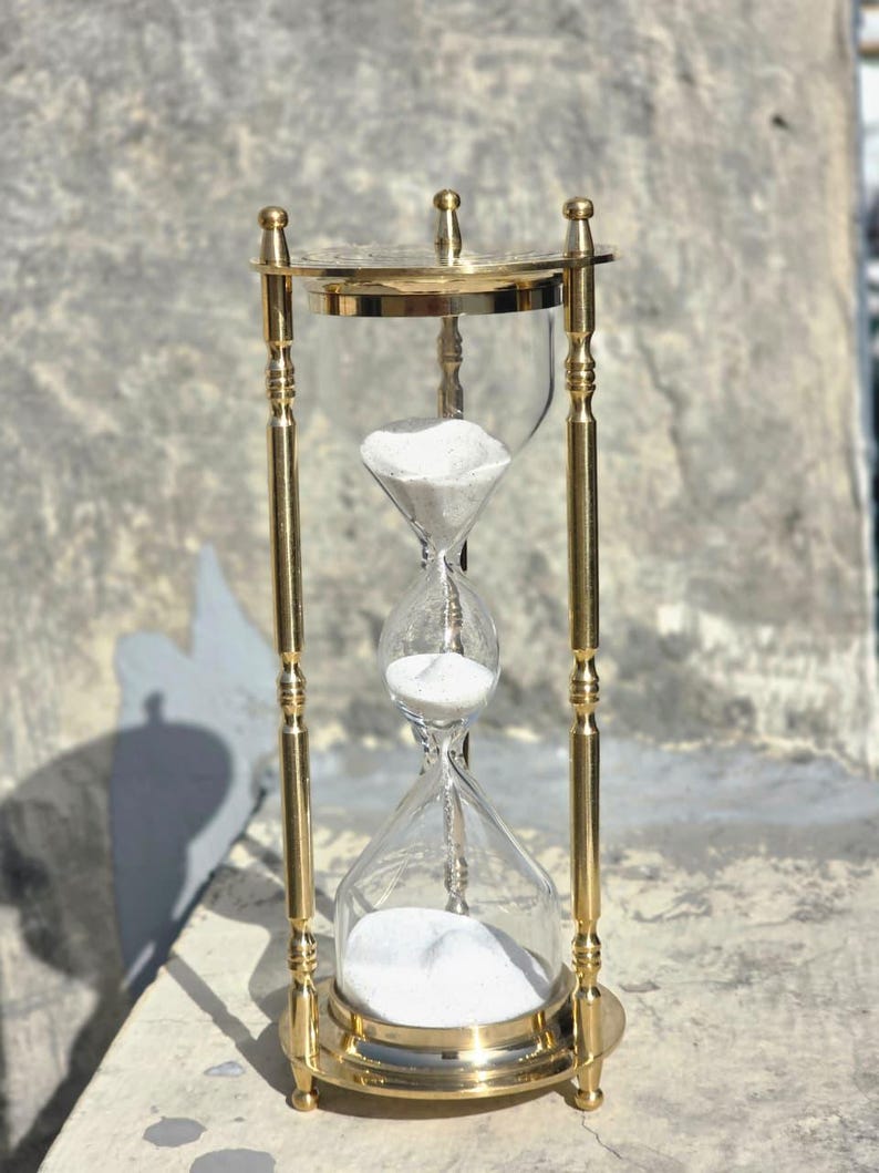 May include: A brass hourglass with clear glass chambers and white sand. The hourglass has a decorative, ornate design with a round base and top. The sand is flowing from the top chamber to the bottom chamber. The hourglass is a classic timekeeping device.