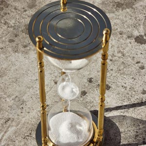 May include: An hourglass with a gold-colored metal frame and a clear glass bulb. The top and bottom of the hourglass are circular, with a series of concentric circles on the top. White sand is flowing from the top bulb to the bottom.