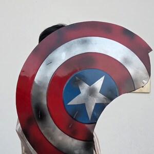 May include: A Captain America shield prop, featuring a red, white, and blue color scheme. The shield has a circular design with a white star on a blue center, surrounded by concentric red and silver rings. The prop is slightly damaged.
