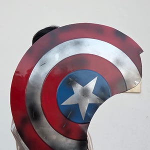 May include: A Captain America shield prop, featuring a red, white, and blue color scheme. The shield has a central blue circle with a white star, surrounded by concentric red and silver bands. The shield is slightly weathered.