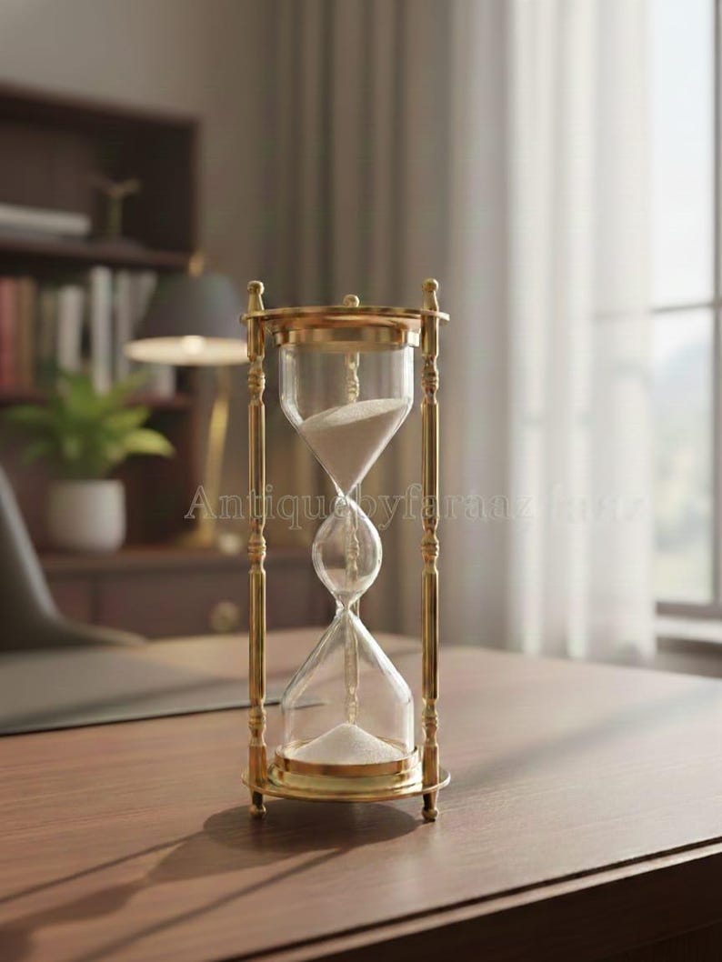 May include: An hourglass with a brass frame and clear glass bulbs. The hourglass is filled with white sand. The frame has decorative vertical bars. The hourglass is sitting on a wooden desk.