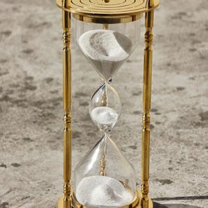 May include: A gold-colored hourglass with clear glass chambers filled with white sand. The hourglass has a decorative, ornate design with a circular top and base. The sand is flowing from the top chamber to the bottom chamber.
