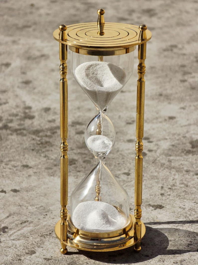 May include: A gold-colored hourglass with clear glass chambers filled with white sand. The hourglass has a decorative, ornate design with a circular top and base. The sand is flowing from the top chamber to the bottom chamber.