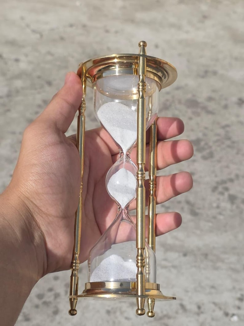 May include: An hourglass with a clear glass body and white sand. The hourglass is framed by a gold-colored metal structure with decorative details. The design is symmetrical, with a base and top.
