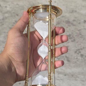 May include: An hourglass with a clear glass body and white sand. The hourglass is framed by a gold-colored metal structure with decorative details. The design is symmetrical, with the sand flowing from the top to the bottom chamber.