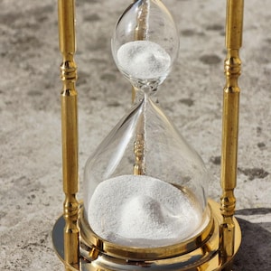 May include: A classic hourglass with a gold-colored metal frame and clear glass bulbs. White sand is visible in the top bulb, slowly flowing to the bottom. The hourglass is a decorative item.