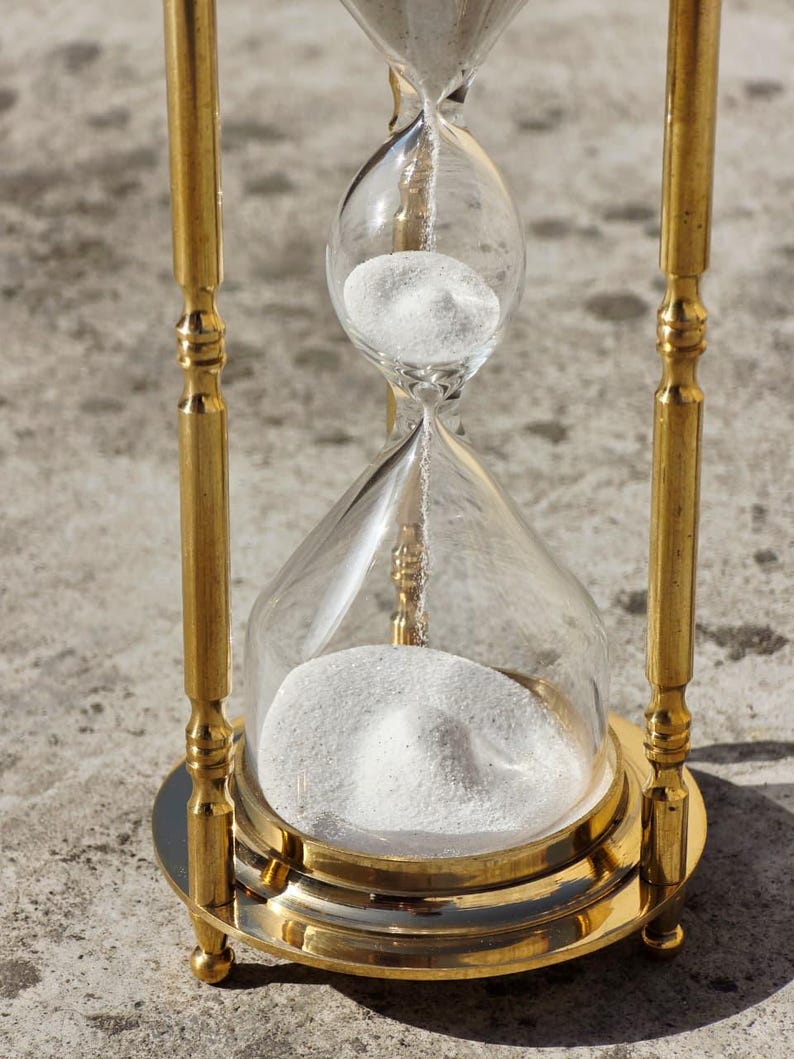May include: A classic hourglass with a gold-colored metal frame and clear glass bulbs. White sand is visible in the top bulb, slowly flowing to the bottom. The hourglass is a decorative item.