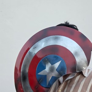 May include: A Captain America shield with a red, white, and blue color scheme. The shield features a central blue circle with a white star, surrounded by concentric rings of silver, white, and red. The shield is a prop or costume accessory.