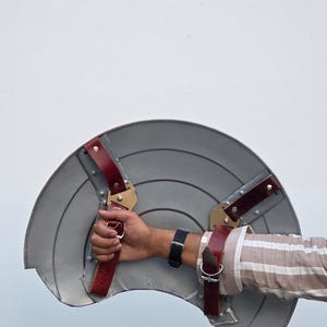 May include: A gray, semi-circular shield with a series of concentric circles. The shield has red leather straps and gold-colored metal brackets for securing the shield to the arm. The shield is held by a person.