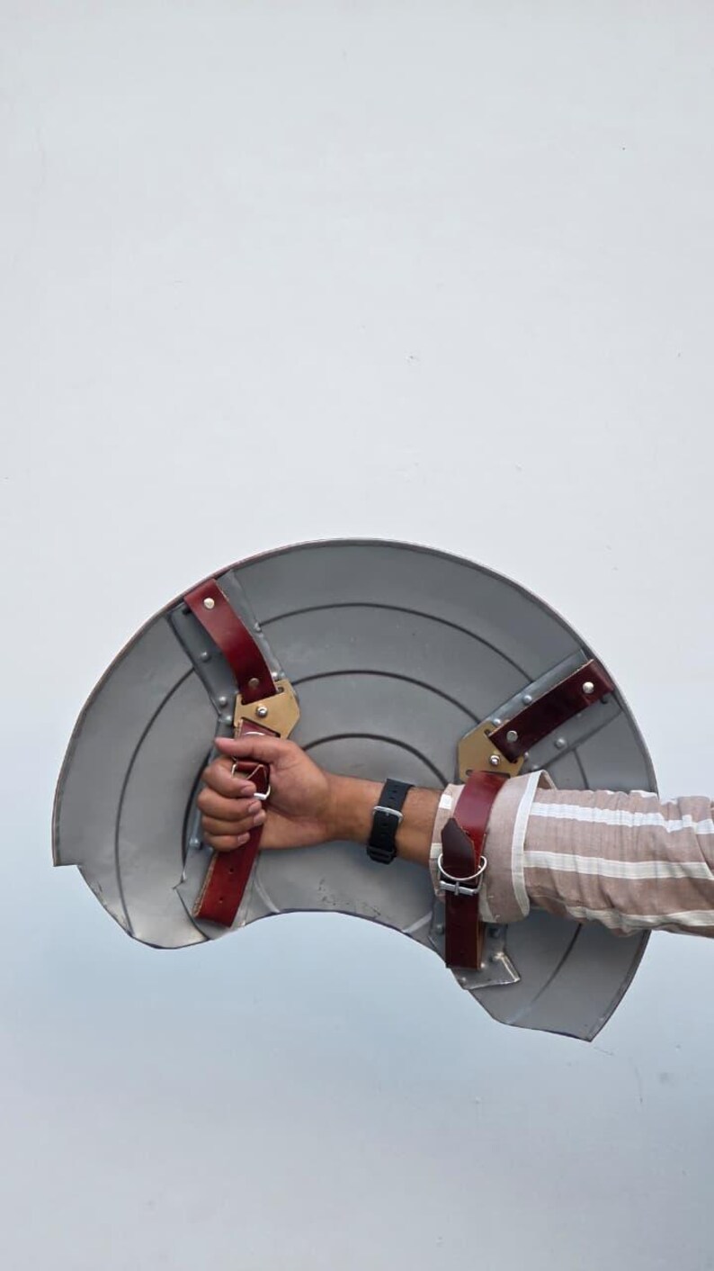 May include: A gray, semi-circular shield with a series of concentric circles. The shield has red leather straps and gold-colored metal brackets for securing the shield to the arm. The shield is held by a person.