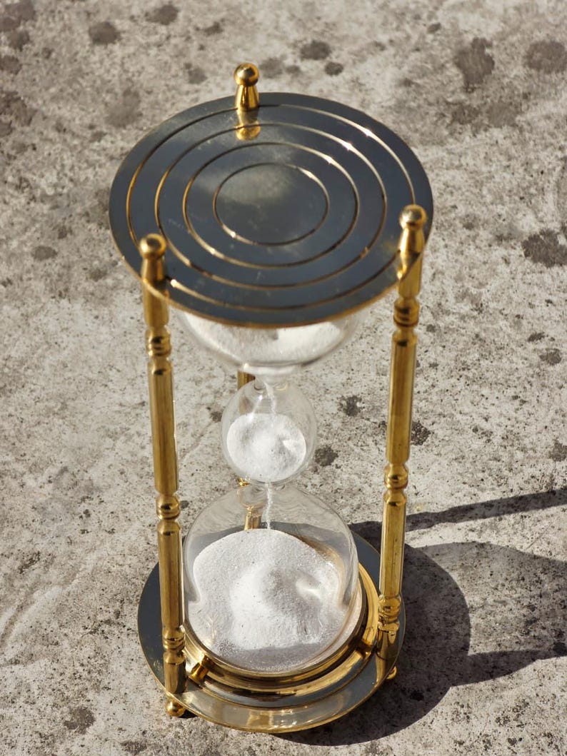 May include: An hourglass with a gold-colored metal frame and a clear glass bulb. The top and bottom of the hourglass are circular, and the sand is white. The hourglass is sitting on a concrete surface.