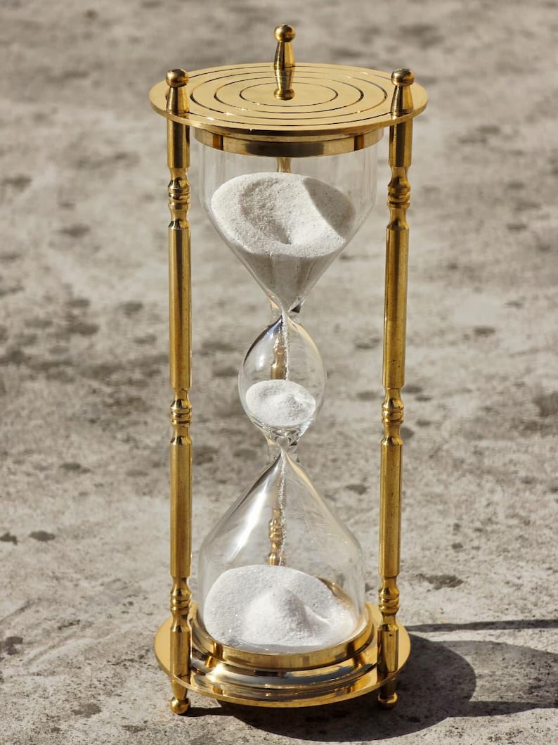May include: A gold-colored hourglass with clear glass chambers filled with white sand. The hourglass has a decorative, ornate design with a circular top and base. The sand is flowing from the top chamber to the bottom chamber.
