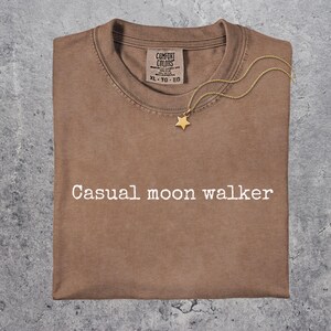 May include: A folded brown t-shirt with the text "Casual moon walker" in white. A gold-colored star pendant necklace rests on the shirt. The shirt has a Comfort Colors tag.