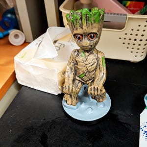 May include: A Groot figurine, featuring a detailed design with brown and green tones. The figure has large, expressive eyes and is posed on a light blue base. The figurine is a collectible item.