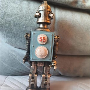 Può includere: Una statuetta di robot in stile vintage con finitura metallica. Il robot ha una piastra pettorale azzurra con il numero "55" e un manometro. La testa del robot ha due occhi rotondi e una parte superiore curva.