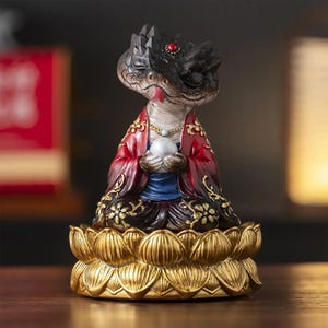 May include: A detailed figurine of a snake-like creature in a seated pose, holding a pearl. The figure wears a red robe with gold accents, a blue undershirt, and is seated on a gold lotus flower base. The snake's head is black with a red jewel.
