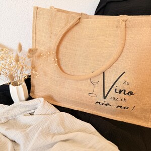 Könnte beinhalten: Eine beigefarbene Jutetasche mit einer Weinglasgrafik und den Worten "Zu Vino sag ich nie no!" in Schwarz. Die Tasche hat zwei Griffe und liegt auf einer schwarzen Oberfläche.