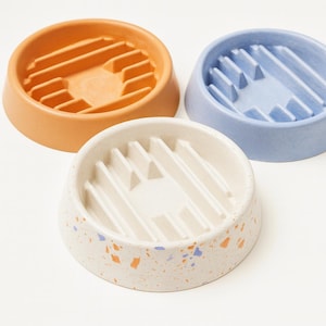 May include: Three ceramic pet bowls in orange, blue, and speckled white. Each bowl has a circular design with raised interior ridges. The bowls are designed to slow down eating for pets.