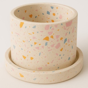 Peut inclure: Pot de fleurs cylindrique avec soucoupe assortie. Le pot en céramique blanc cassé est parsemé d'éclats de terrazzo colorés roses, jaunes et bleus. Le pot a un bord lisse et repose sur une soucoupe assortie.