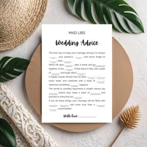 May include: A white Mad Libs wedding advice card with black text, featuring fill-in-the-blank prompts. The card is on a brown surface, surrounded by decorative elements like a woven basket and greenery.