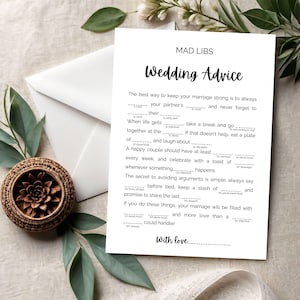 May include: A white Mad Libs wedding advice card with black text, a white envelope, and a woven basket with a decorative flower. The card reads "Wedding Advice" and includes fill-in-the-blank prompts.
