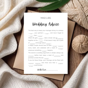 May include: A white Mad Libs wedding advice card with black text, including fill-in-the-blank prompts. The card is on a wooden surface, with a beige envelope and decorative yarn in the background.
