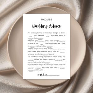 May include: A white Mad Libs wedding advice card with black text, including fill-in-the-blank prompts. The card is on a beige plate, with the words "Wedding Advice" in elegant script.