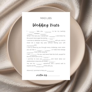 May include: A white Mad Libs Wedding Vows sheet on a beige plate. The text includes fill-in-the-blank prompts for vows, such as "I promise to dedicate my time to..." and "I promise to love you..." The sheet is printed with black ink.