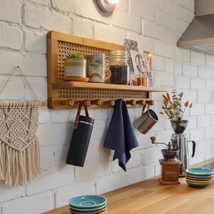 Puede incluir: Una estanter&iacute;a de pared de bamb&uacute; con respaldo de ca&ntilde;a tejida, que contiene una planta en maceta, tazas y un tarro. Debajo, una fila de ganchos sostiene un termo negro, una toalla azul y una taza de metal. Tambi&eacute;n se ve un tapiz de macram&eacute; y una pila de platos coloridos.